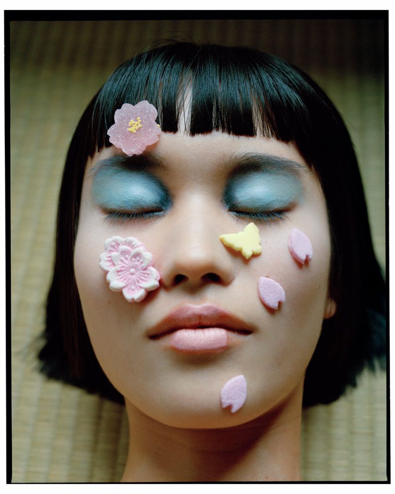 Portrait of Asian woman with closed eyes, face adorned with flower and butterfly candy against a woven mat background in soft lighting.
