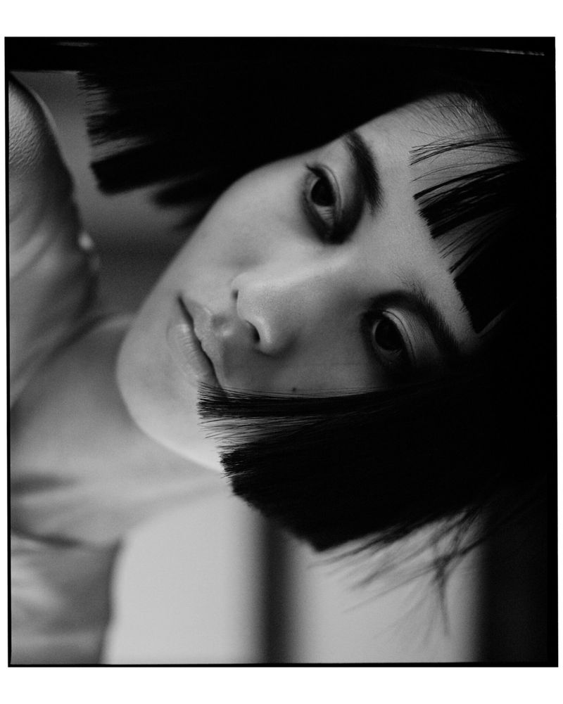 Close-up black and white portrait of a woman with a sharp blunt black bob and micro bangs looking toward the camera.
