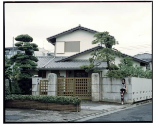 Model in a patterned top and cutout leggings leaning against an exterior concrete wall of a traditional Japanese house.