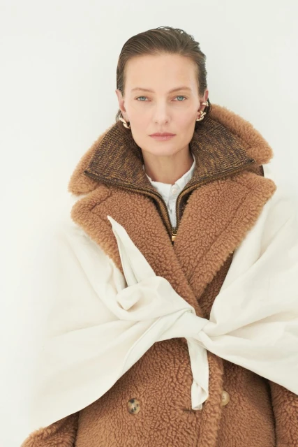 A poised woman wearing a brown teddy-bear coat and statement gold earrings against a minimalist white backdrop in a portrait-style fashion shot.
