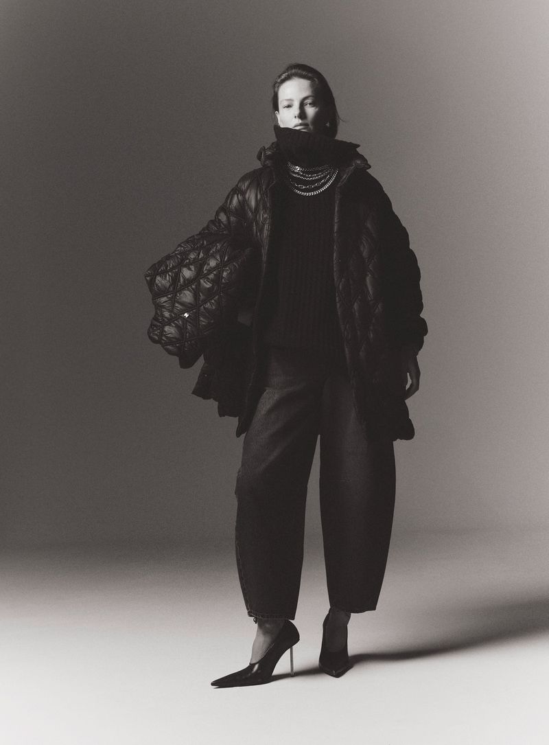 Fashion photo of woman in oversized quilted coat, dark turtleneck and balloon pants, plus heels, pearls, and neutral backdrop.