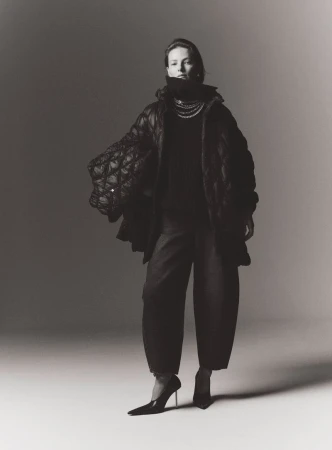 Fashion photo of woman in oversized quilted coat, dark turtleneck and balloon pants, plus heels, pearls, and neutral backdrop.