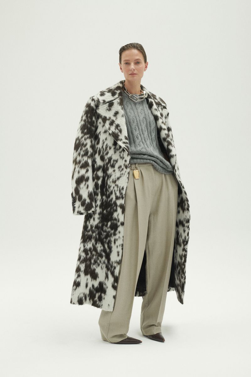 Full-length shot of a female fashion model wearing a black and white coat, gray sweater, and tan pants against a white backdrop.