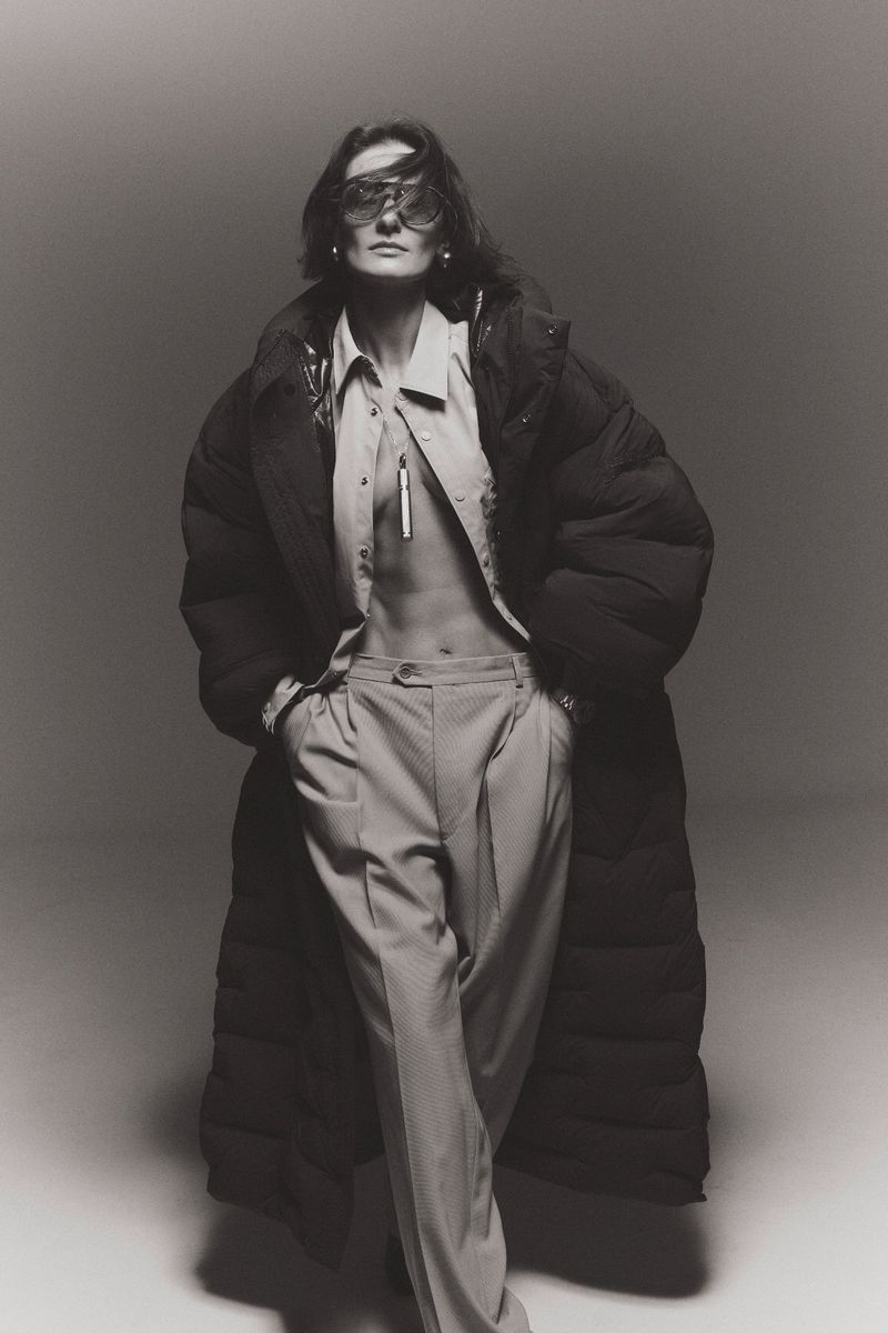 Monochrome fashion portrait of adult female in an oversized puffer coat and eyewear, standing in a light grey studio setting.