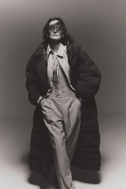 Monochrome fashion portrait of adult female in an oversized puffer coat and eyewear, standing in a light grey studio setting.