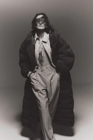 Monochrome fashion portrait of adult female in an oversized puffer coat and eyewear, standing in a light grey studio setting.