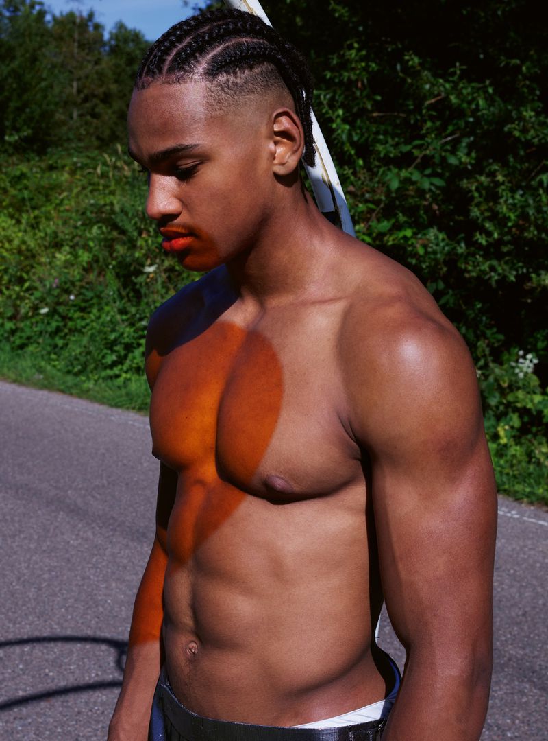 Shirtless male model poses outdoors with cornrow braids, showcasing athletic build against a green tree and gray road background.