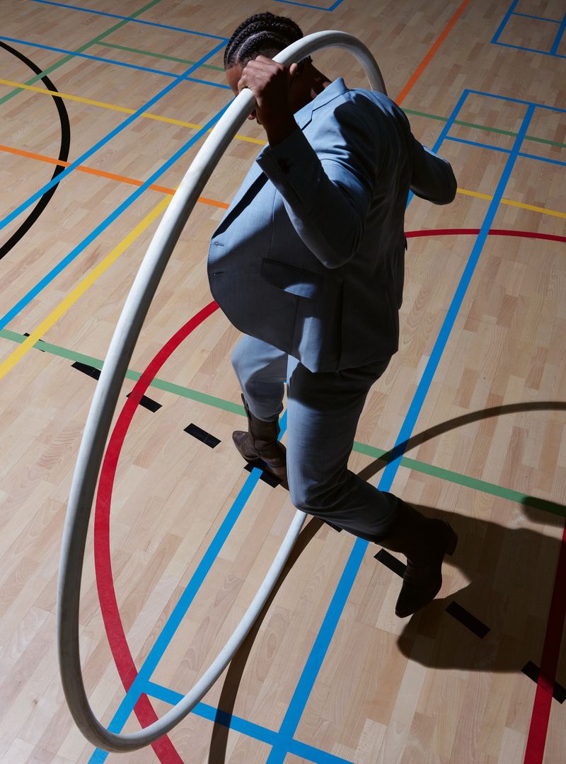 Stylish adult male in a light blue suit steps through a white hula hoop on a colourful indoor court with natural light and shadows.
