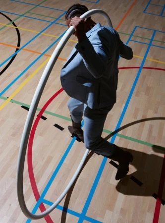 Stylish adult male in a light blue suit steps through a white hula hoop on a colourful indoor court with natural light and shadows.