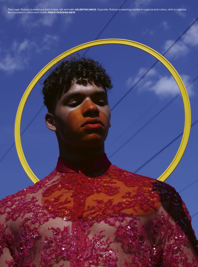 Man with orange face paint wears red lace top in bright outdoor fashion shot against blue sky with yellow halo prop.