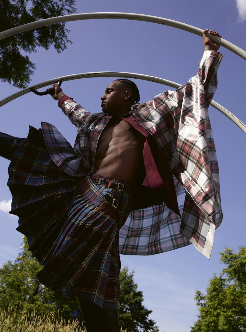 An adult male poses in a plaid kilt and jacket while holding onto a metal structure in an outdoor park setting on a sunny day.