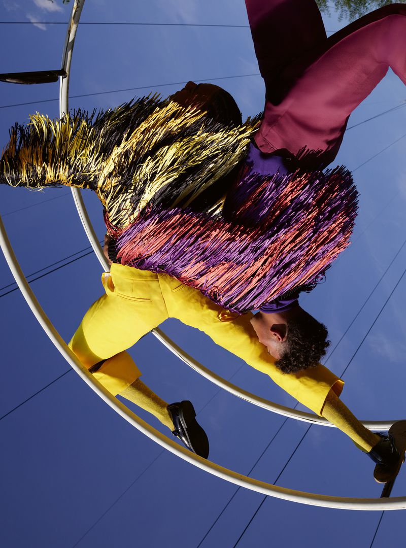 Two fashion models in yellow and pink outfits, climbing on a metal circular structure against a blue sky with power lines.