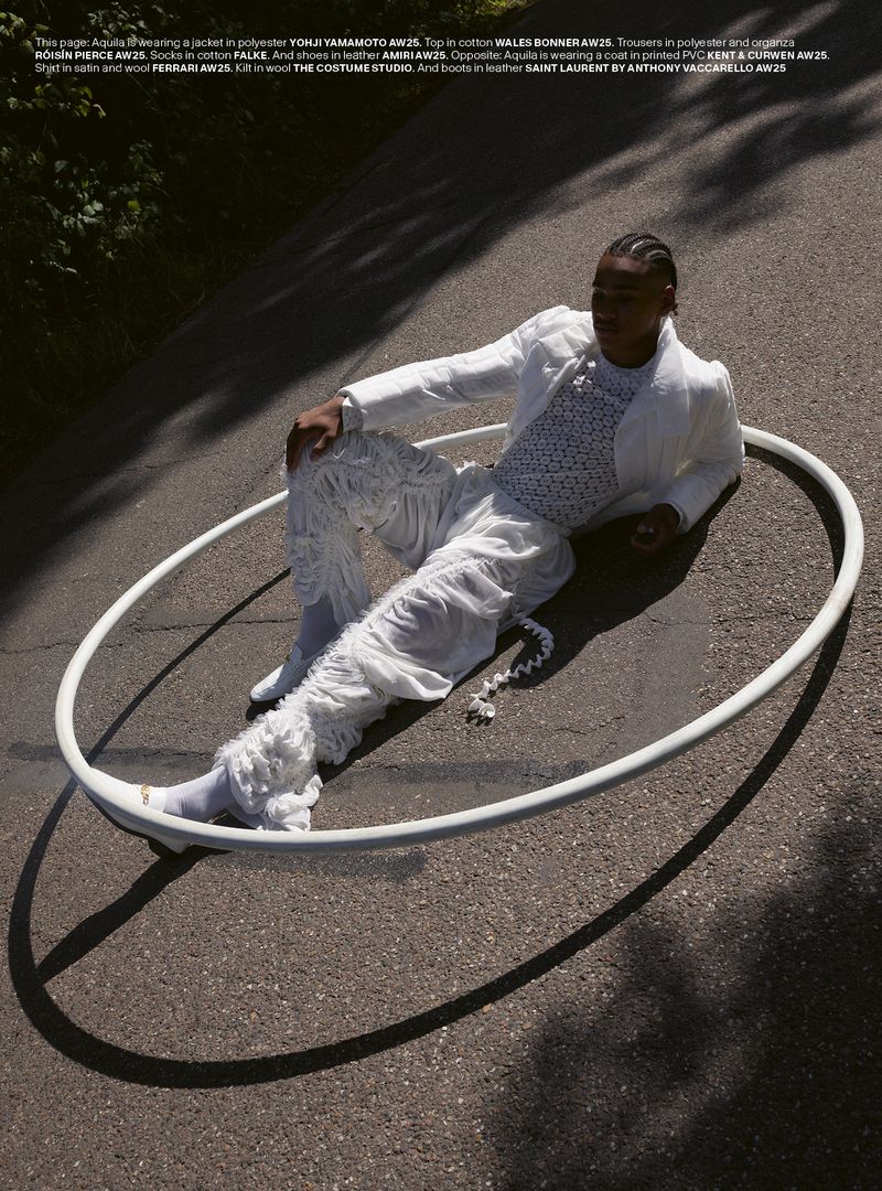 A young adult male poses outdoors in a white jacket, patterned top, and textured pants, seated within a white circular frame.