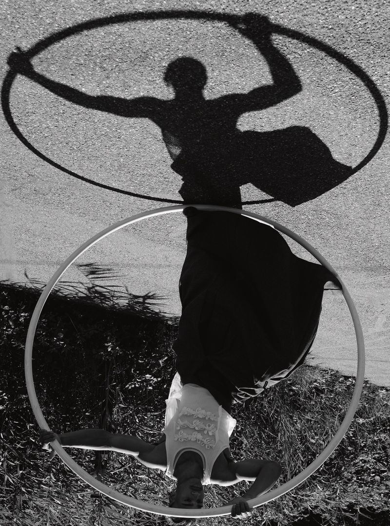Monochromatic outdoor shot of person holding hoop with arms extended while upside down, shadow visible on pavement surface.