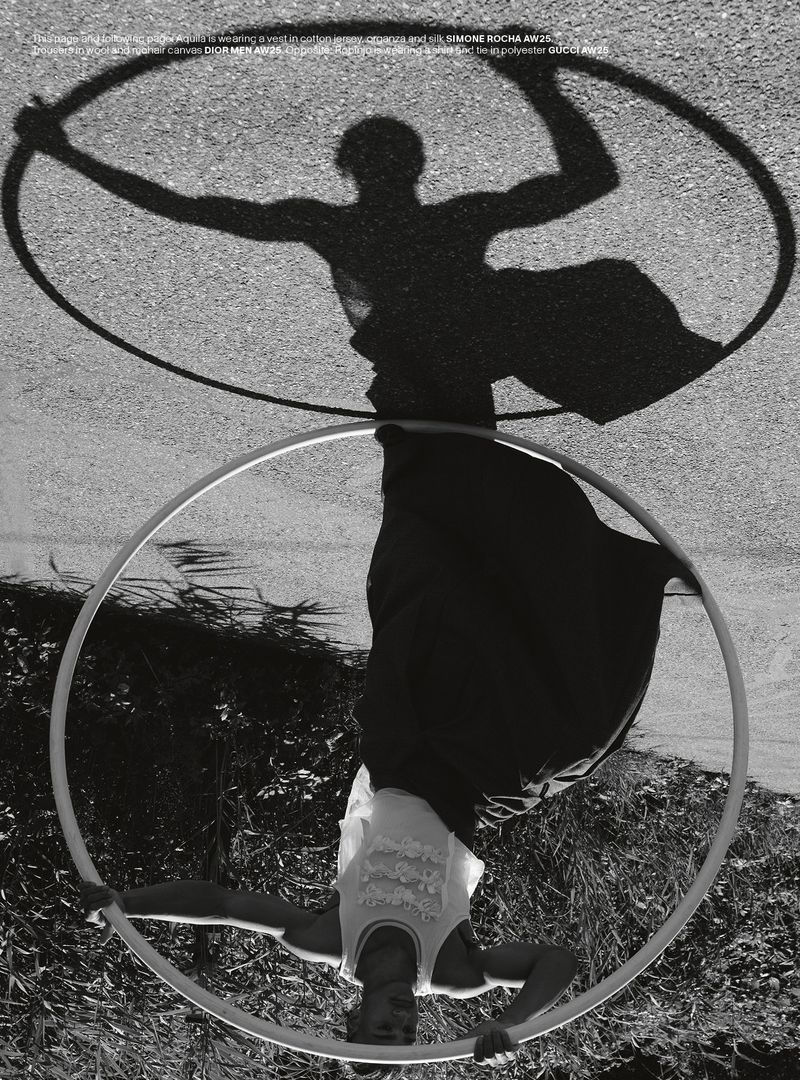 A black and white fashion shot presents an adult female in a white top and dark pants performing a hoop routine outdoors with shadows.