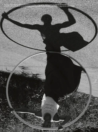 A black and white fashion shot presents an adult female in a white top and dark pants performing a hoop routine outdoors with shadows.