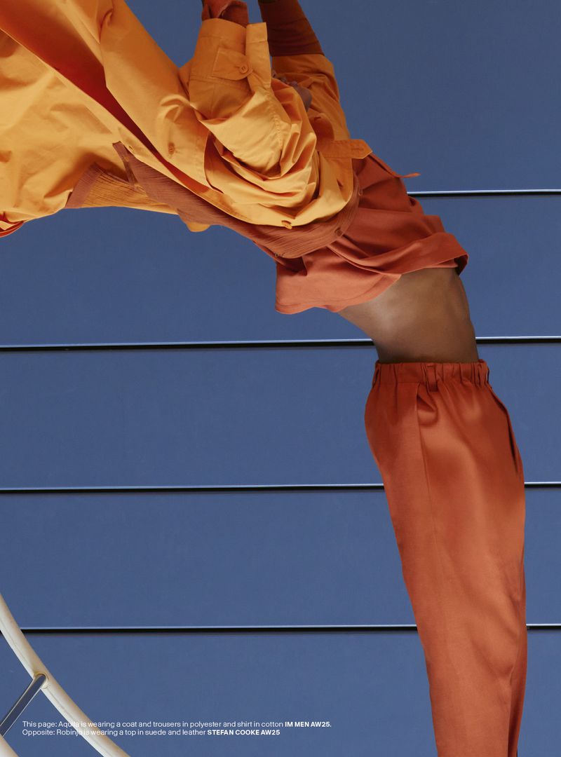 Fashion editorial shot: A model in an orange coat and trousers poses against a blue, slatted wall with white steps for a striking contrast.