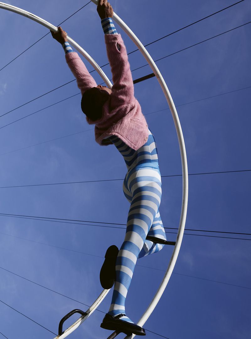 Person in blue and white striped leggings, pink sweater, and black shoes poses on white metal structure against a bright blue sky.