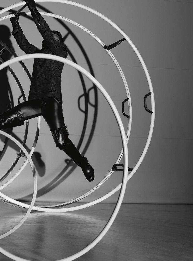 Monochrome fashion shot of a person in a black suit and leather pants jumping through several white circular metal structures.