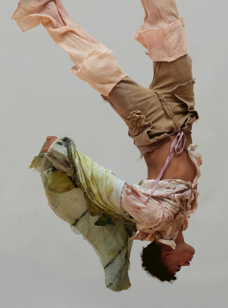 Upside-down view of a person with curly hair wearing patchwork-style clothing and a pink ribbon against a solid white background.