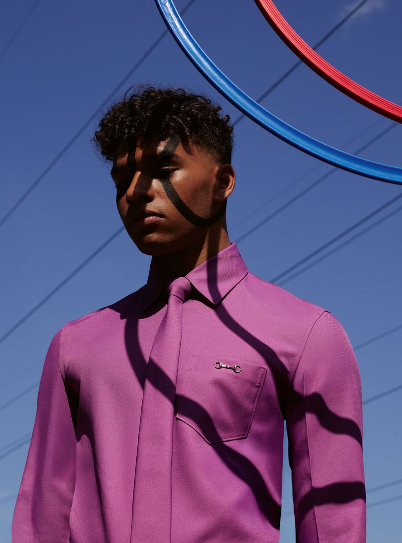 Young man with dark hair and purple shirt posing outdoors against a blue sky with geometric colorful circles and overhead power lines.