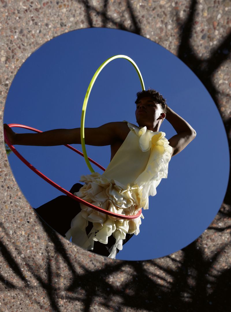 A person in a unique top posing in a circular mirror with a blue sky reflecting in the glass, and holding hula hoops.