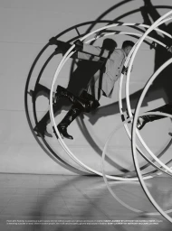 Man wearing a tailored jacket, gloves, and tall boots jumping through a large white hoop structure casting dramatic shadows.