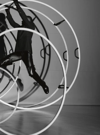 A person in dark tailored clothing and leather boots maneuvers inside a large white circular acrobatic wheel in a monochrome studio.