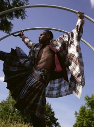 A Black male model wearing a plaid kilt and oversized checked cape holding onto curved metal arch structures outdoors against a blue sky.