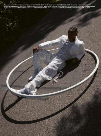 Model in an all-white textured outfit reclining inside a large white hoop resting on an asphalt road in direct sunlight.