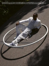 Model in an all-white textured outfit reclining inside a large white hoop resting on an asphalt road in direct sunlight.