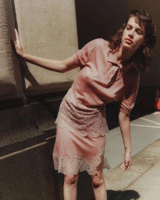 Adult female model in a pink collared shirt and skirt poses near a concrete pillar in soft, natural light; fashion editorial photography.
