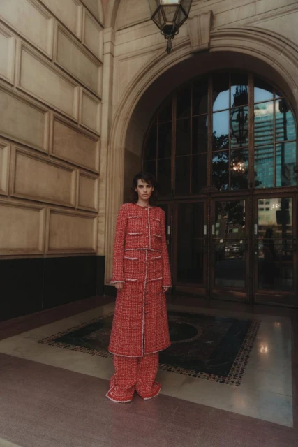 A fashionably dressed woman in a red and white patterned coat and pants stands in front of a grand building with large arched windows.