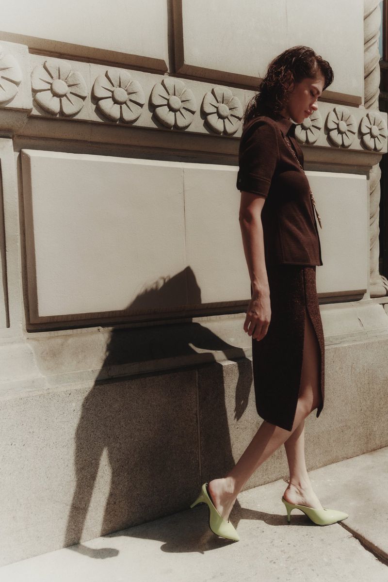 A woman in a brown skirt suit and light green heels poses outdoors beside a stone wall with decorative carvings on a sunny day.