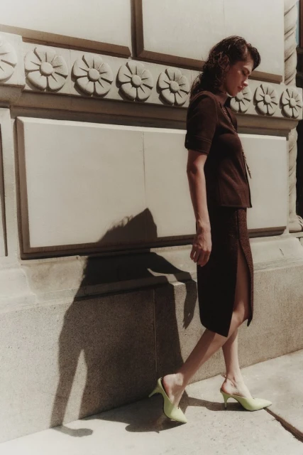 A woman in a brown skirt suit and light green heels poses outdoors beside a stone wall with decorative carvings on a sunny day.