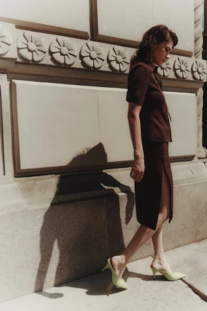 A woman in a brown skirt suit and light green heels poses outdoors beside a stone wall with decorative carvings on a sunny day.