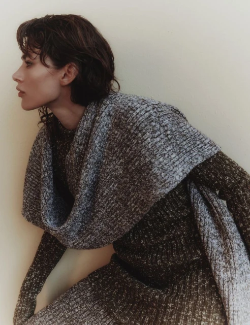 A young adult female with short brown hair poses indoors wearing a patterned sweater and scarf in a studio setting with neutral lighting.