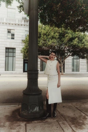 A woman in a light-colored two-piece outfit poses next to a black lamppost on a city street with buildings and trees in the background.