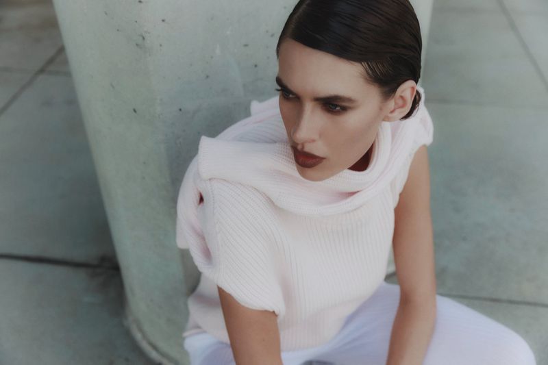 Woman with dark hair poses in a light pink sweater and white pants. Gray concrete provides an urban backdrop.