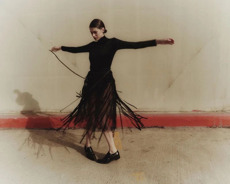Young woman in black turtleneck and fringe skirt dances in photo studio with concrete wall and floor, arms outstretched, motion blur.
