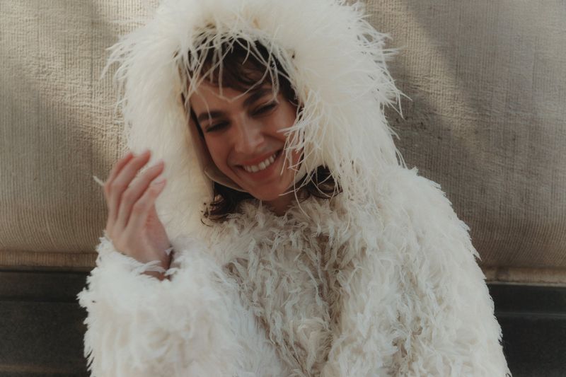 Smiling woman in a white, feathery hooded coat sits outdoors with a grey textured wall behind her. Light, airy fashion photography.