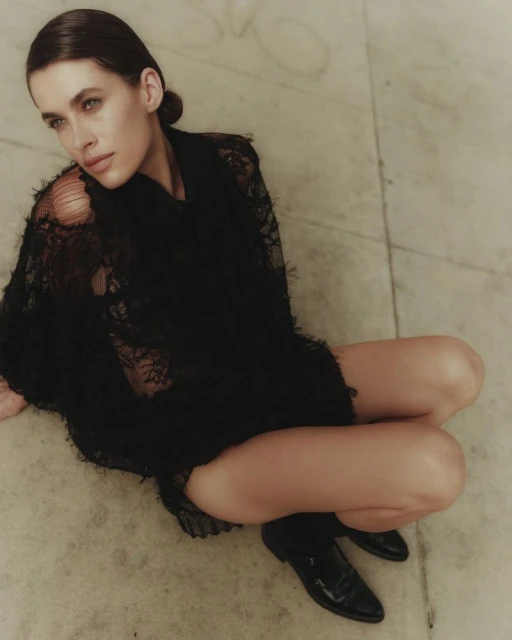 Woman in black lace top and boots sits on a marble floor looking to the left in a studio fashion editorial portrait.
