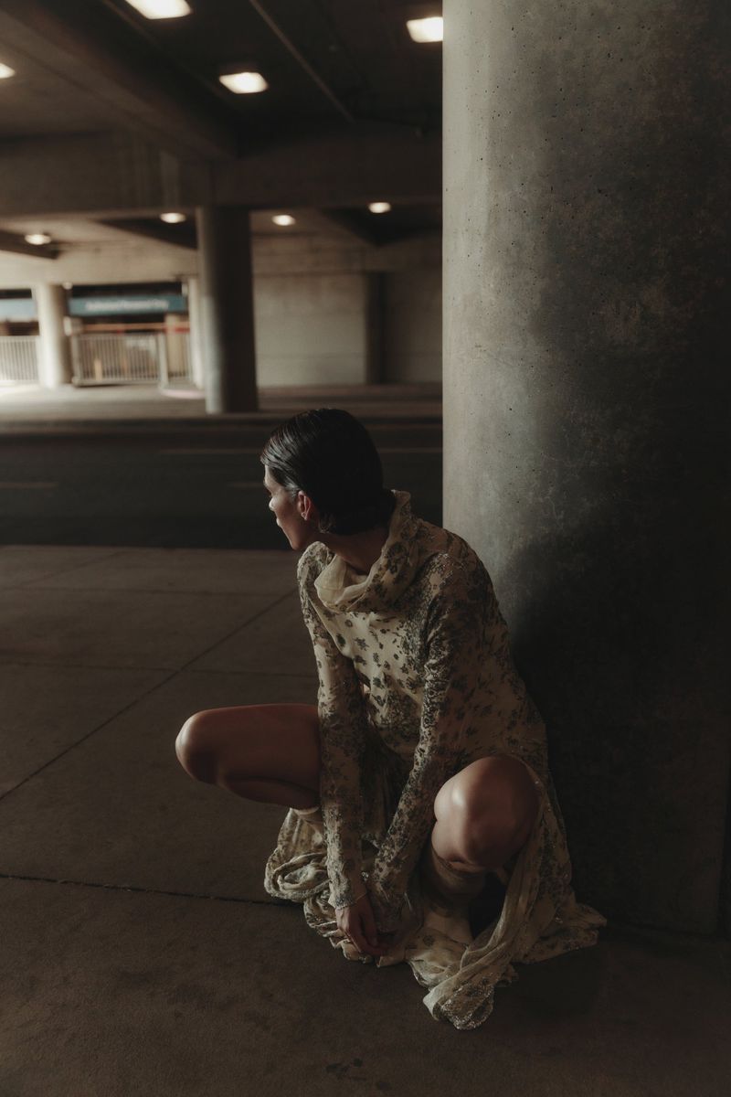 A woman in a patterned dress squats near a concrete pillar in an urban setting with overhead lighting, creating a fashion editorial image.
