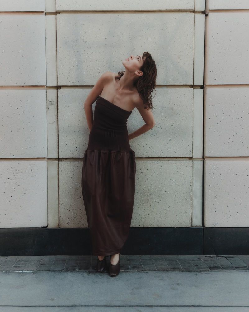 A brunette woman in a brown dress poses against a concrete wall looking upward. The photo is a full shot and well-lit.