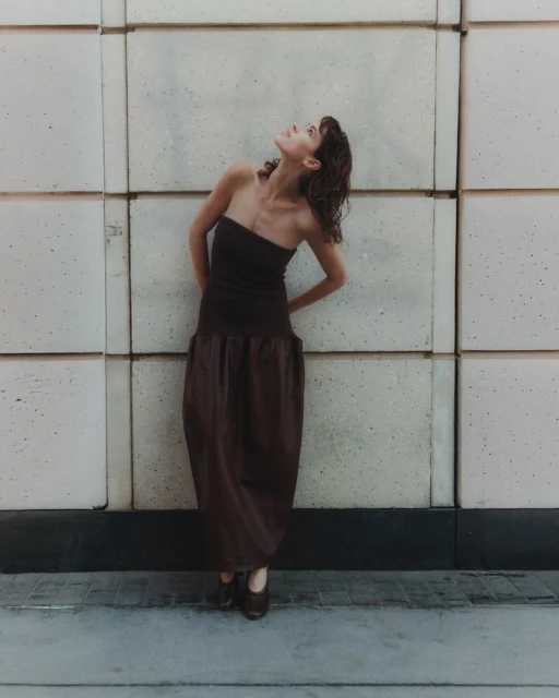 A brunette woman in a brown dress poses against a concrete wall looking upward. The photo is a full shot and well-lit.