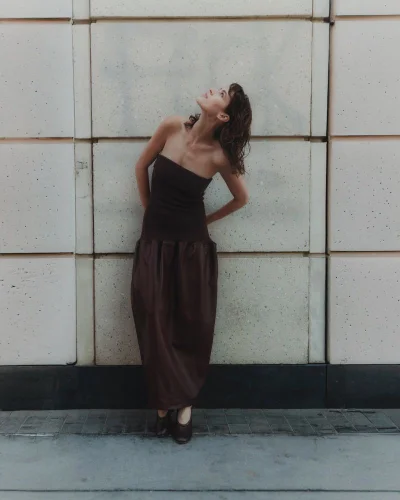 Model Keltie Straith wearing a strapless brown maxi dress leaning against a paneled wall looking upward.