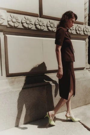 Model Keltie Straith walking past a stone wall in a dark brown two-piece skirt set and lime green pointed mule heels.