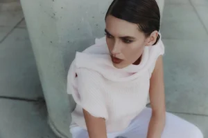 Model Keltie Straith seated in a pale blush sleeveless knit top and white trousers beside a concrete pillar.