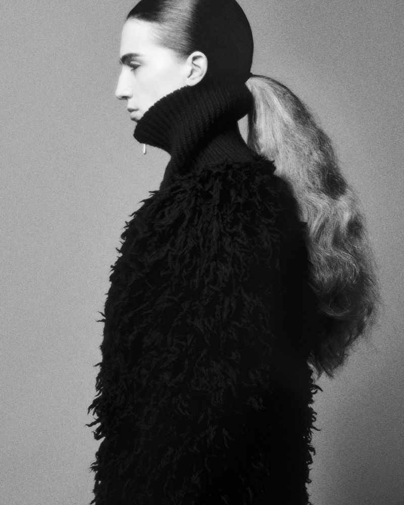 Monochrome fashion portrait: a female model with a dramatic ponytail and a textured, black turtleneck sweater poses in profile.