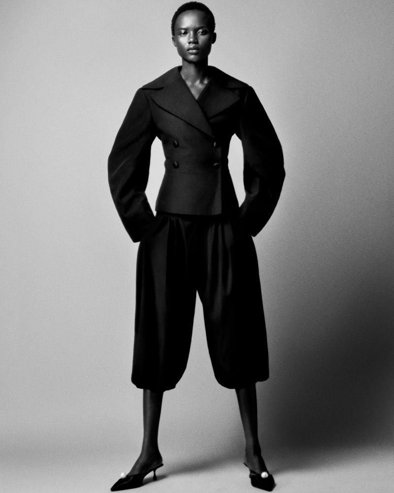 A black and white full shot of a slender Black woman in a dark suit jacket and wide-leg cropped pants against a grey studio backdrop.